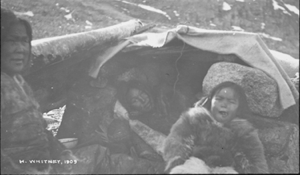 Image of Inuit women and child sit by rock/tarp shelter